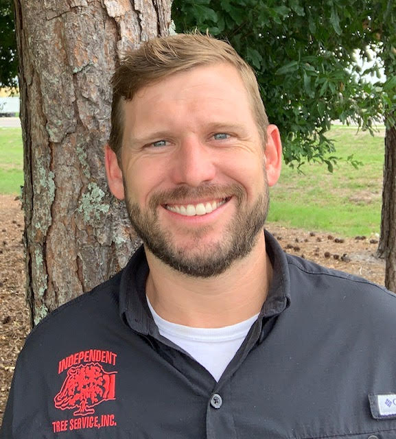 A person with a beard smiles outdoors, standing near a tree. The shirt reads "Independent Tree Service, Inc." There are no landmarks or historical buildings.