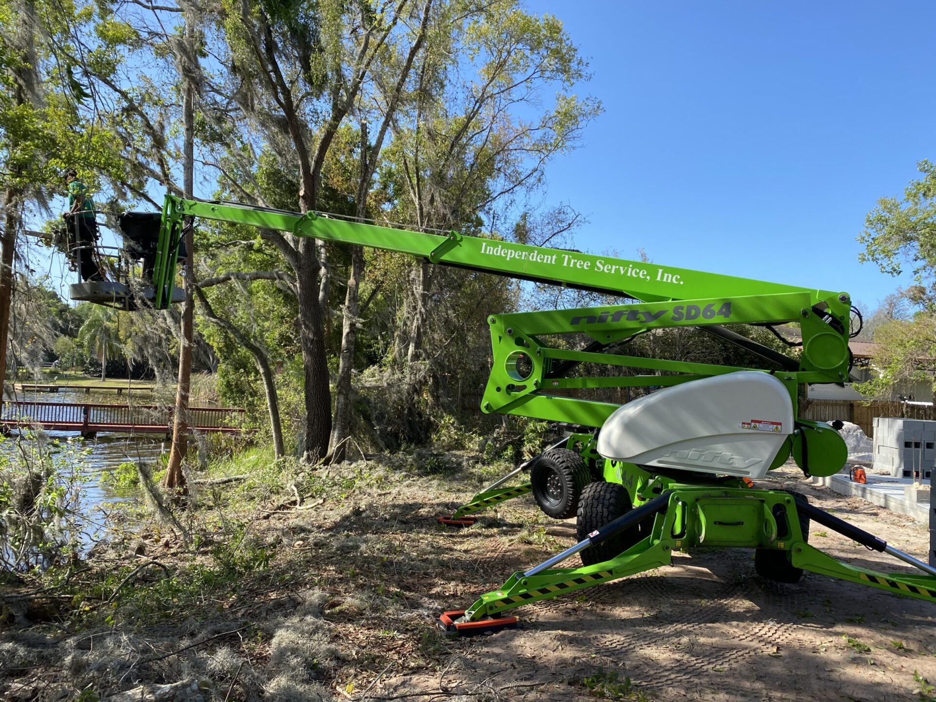 A green tree service lift is extended toward trees by a lake. A person operates it among branches, surrounded by a forested area.