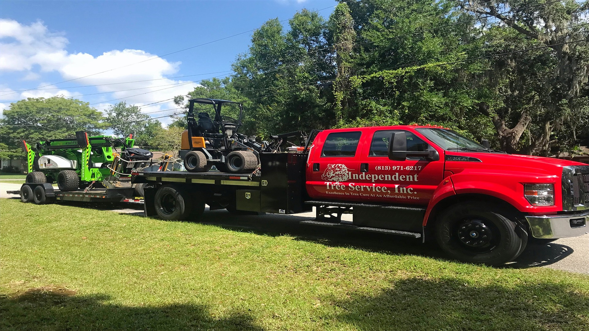 A red truck from Independent Tree Service Inc. transports tree-cutting equipment on a sunny day, surrounded by lush greenery and trees.