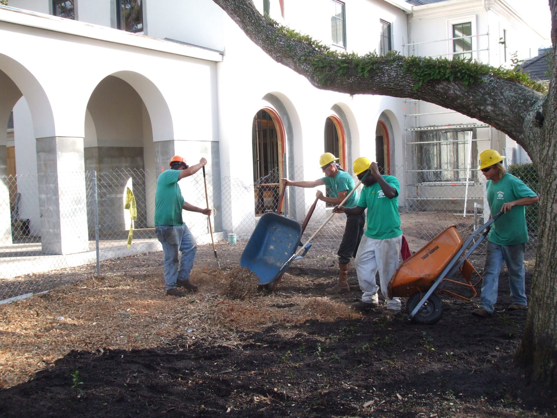 People wearing helmets are landscaping with wheelbarrows and shovels near an arched building facade. Trees and mulch are visible in the foreground.