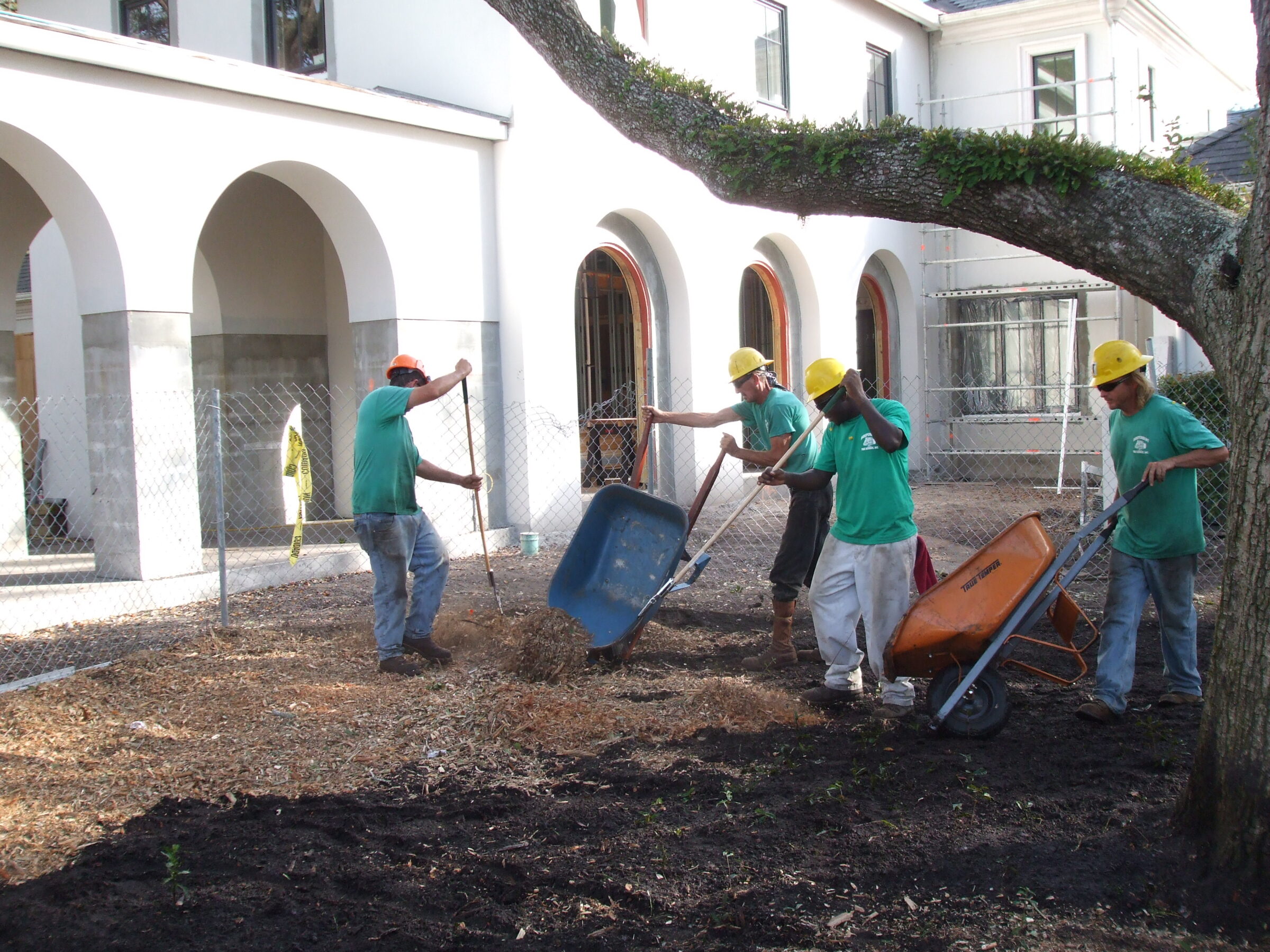 People wearing helmets are landscaping with wheelbarrows and shovels near an arched building facade. Trees and mulch are visible in the foreground.
