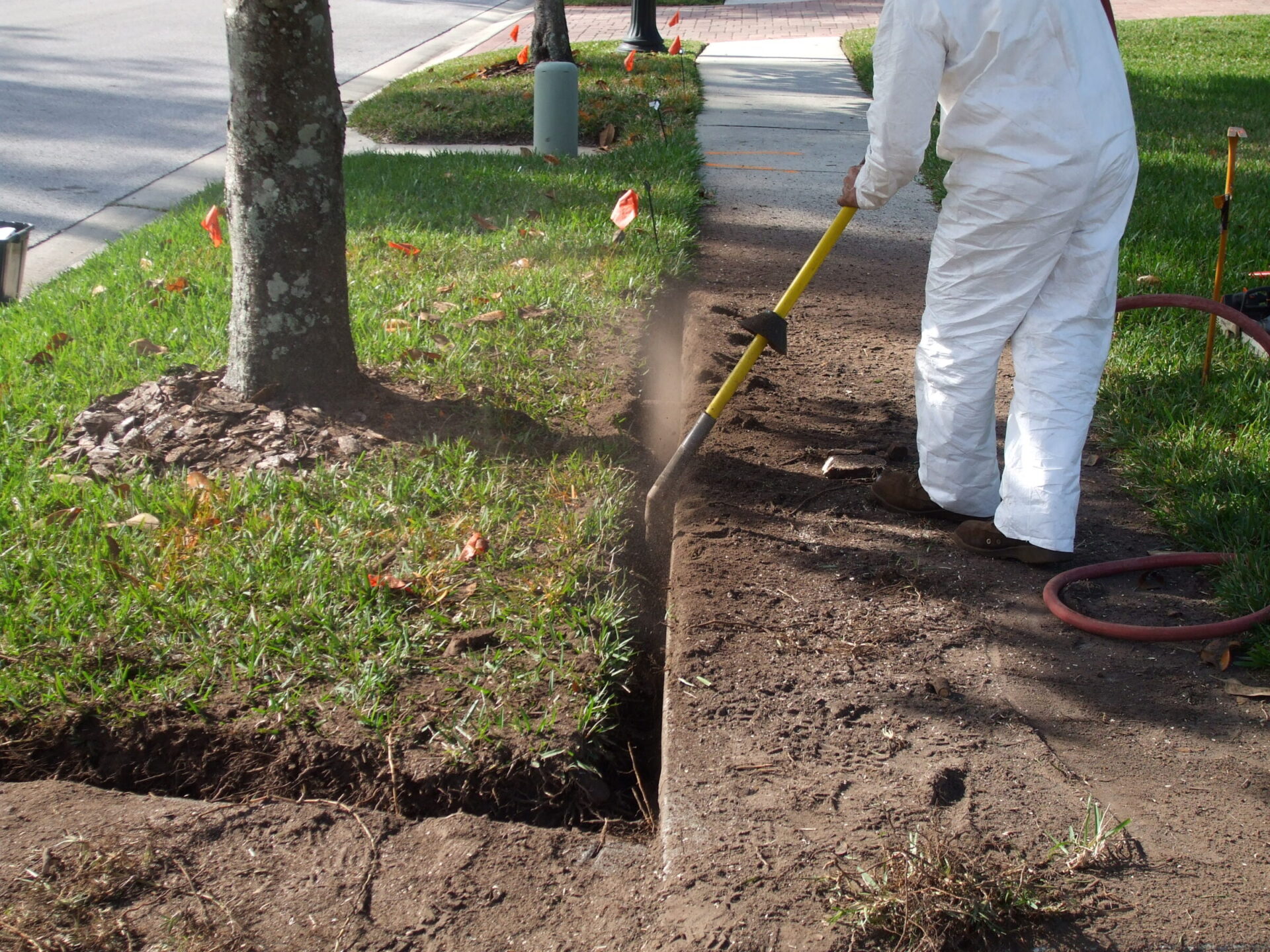 A person in protective gear operates a trenching tool on a grassy sidewalk edge, with orange markers visible nearby.