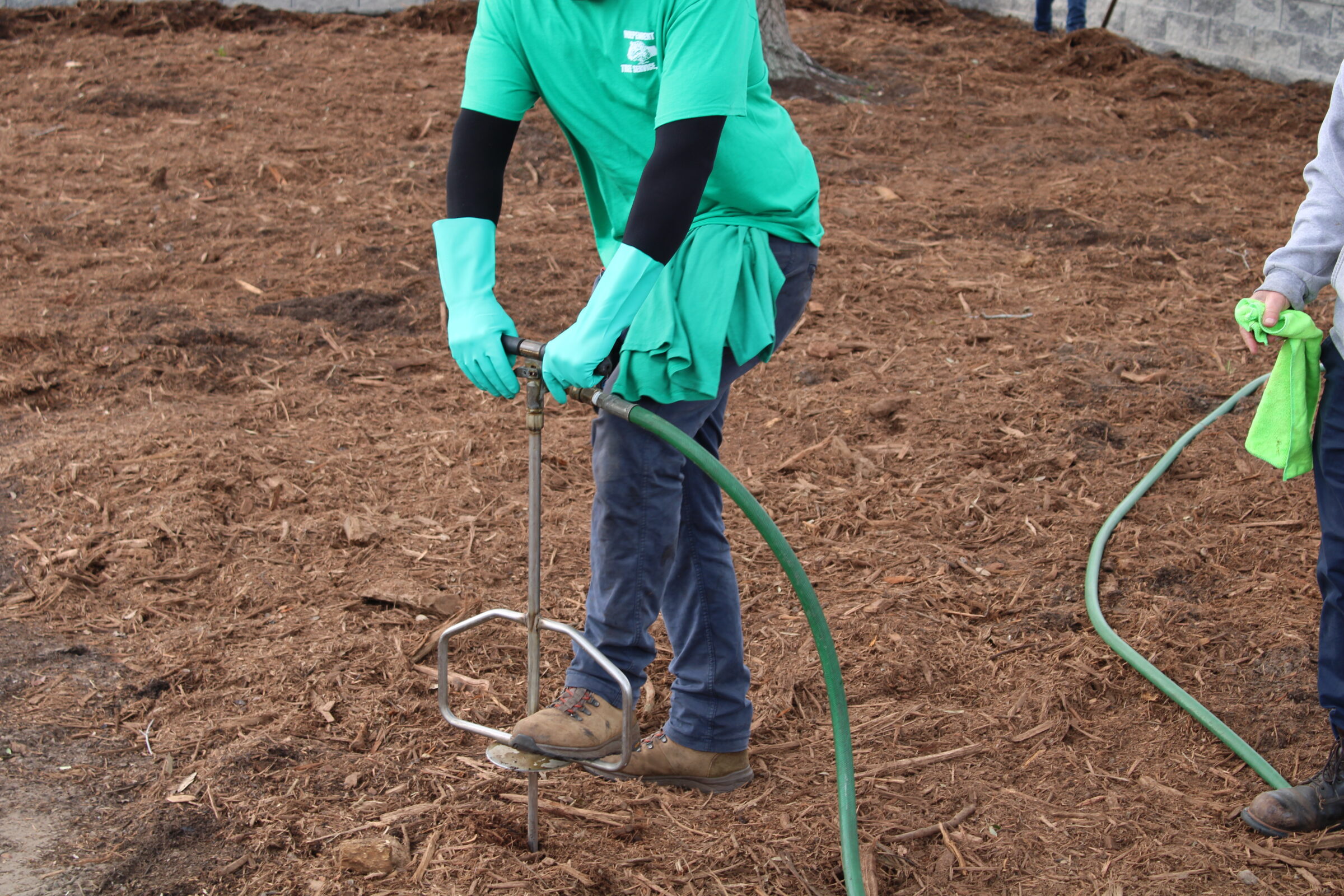 Two people in green attire and gloves are working with a hose and tool on a mulched ground surface, focusing on a task.