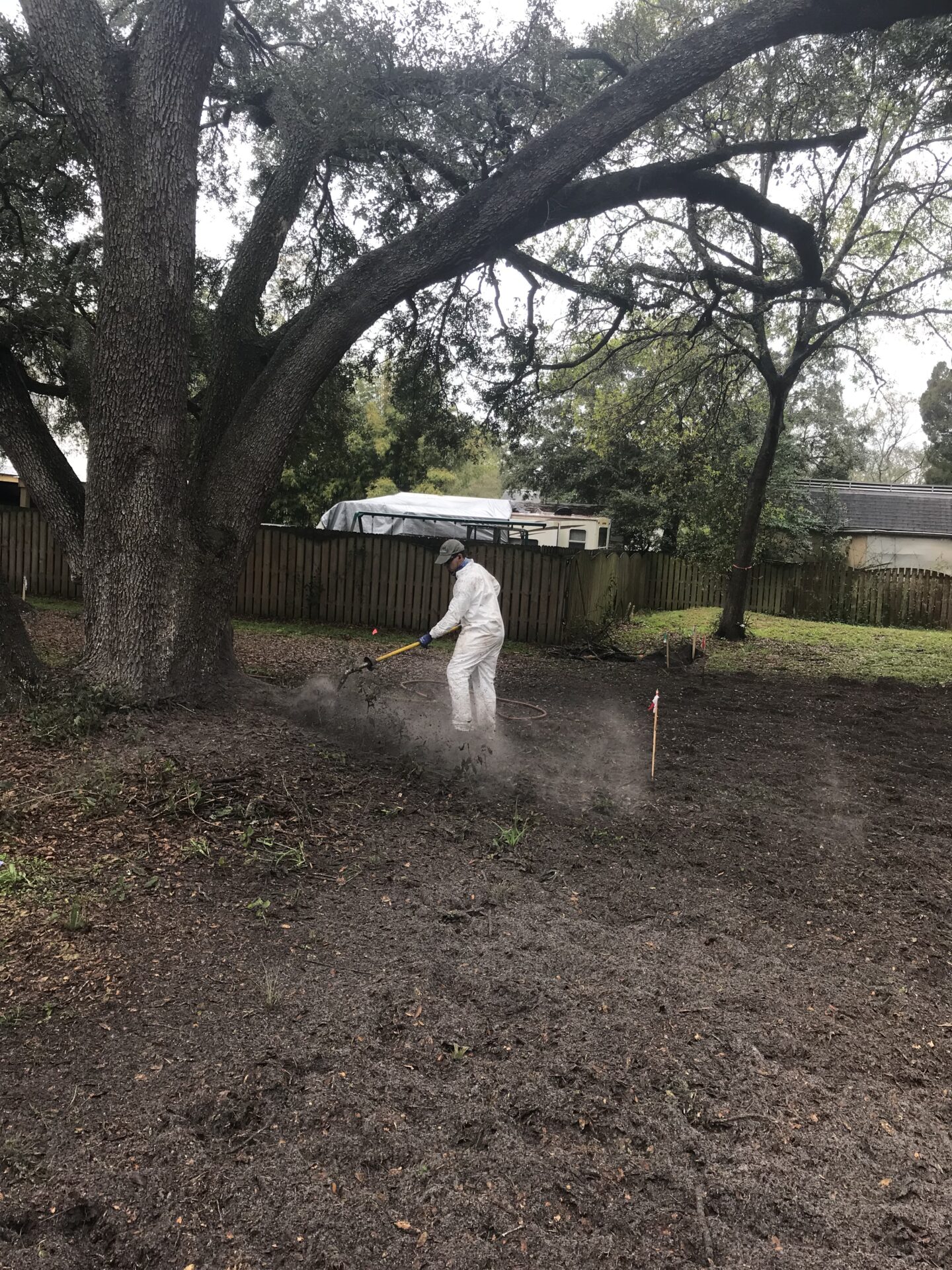 A person in protective gear uses equipment near a large tree in a yard. Wooden fence and trees are visible in the background.