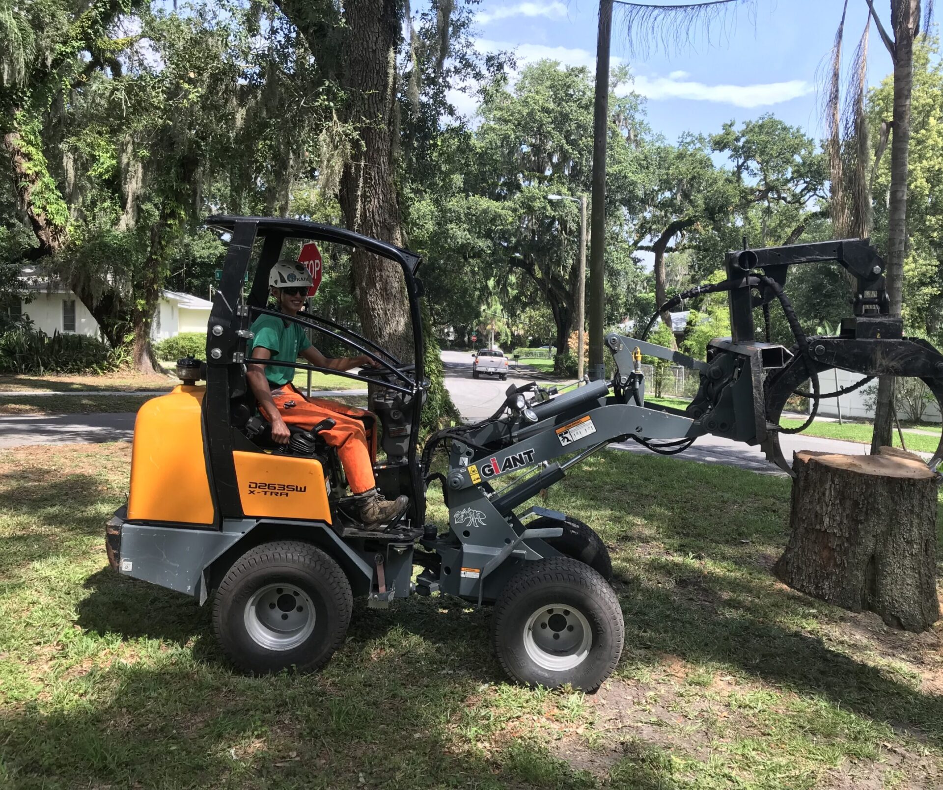 A person operates a mini loader near a tree stump on a suburban street, surrounded by lush greenery and residential homes.