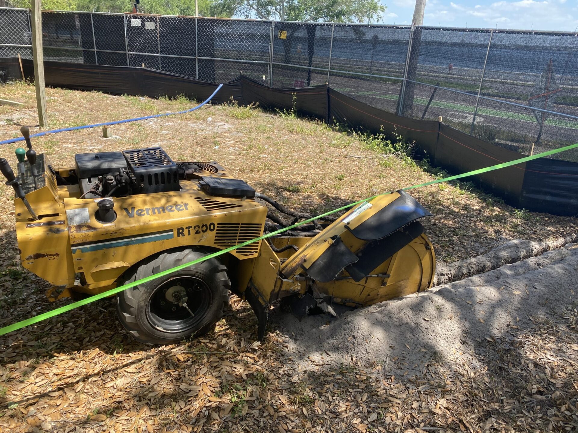 A yellow Vermeer RT200 trencher is parked on a dirt patch, surrounded by fencing and lined with green tape, under a clear sky.