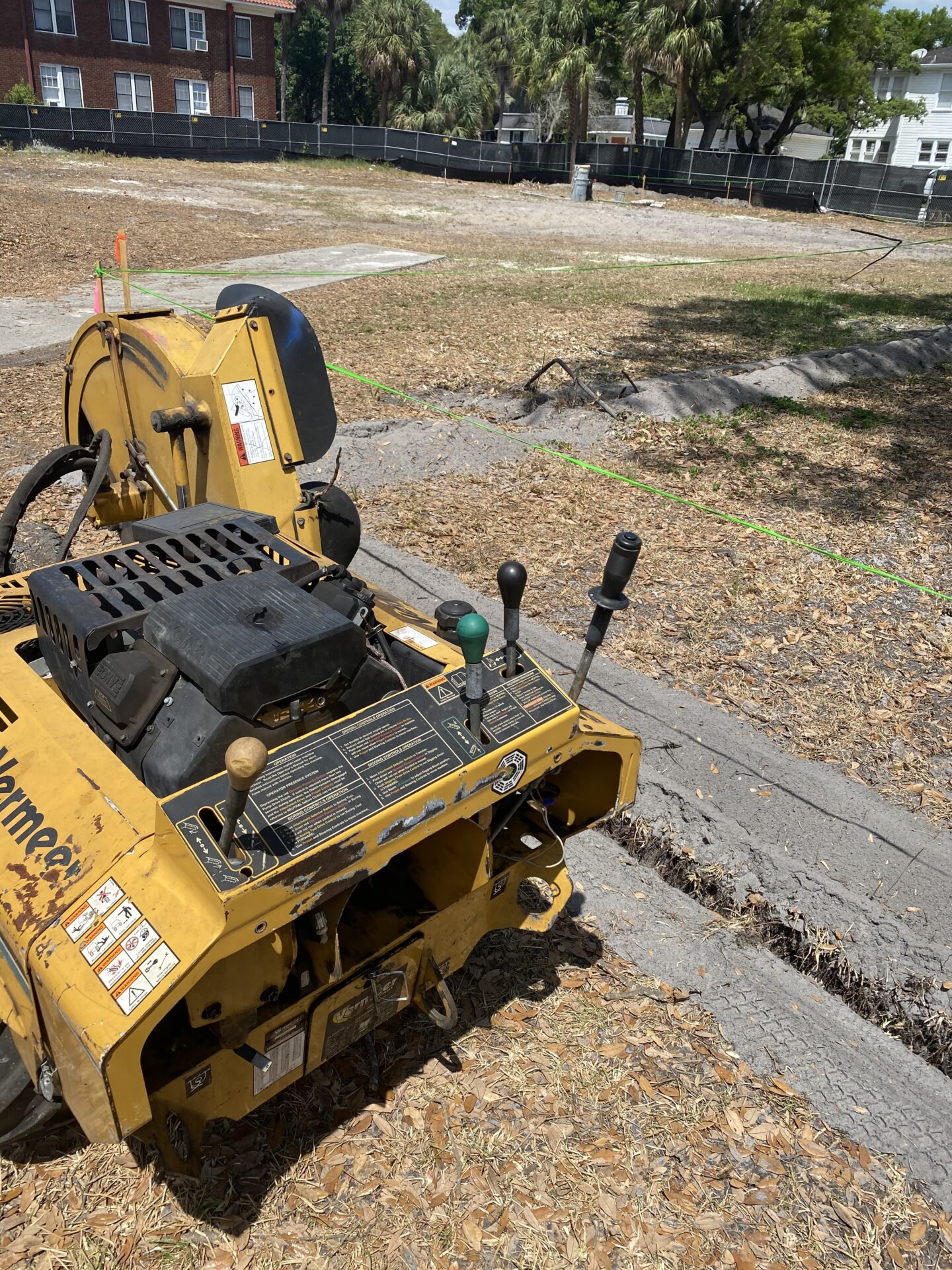 A yellow stump grinder sits on a grassy area near a fenced-off section, with residential buildings and palm trees in the background.