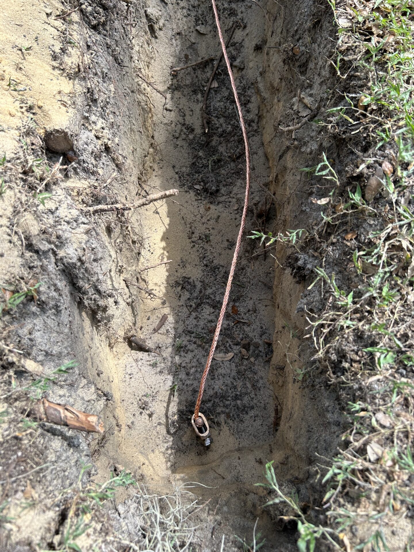 A narrow dirt trench with a copper cable running inside, surrounded by grass and soil, possibly for an underground utility installation.