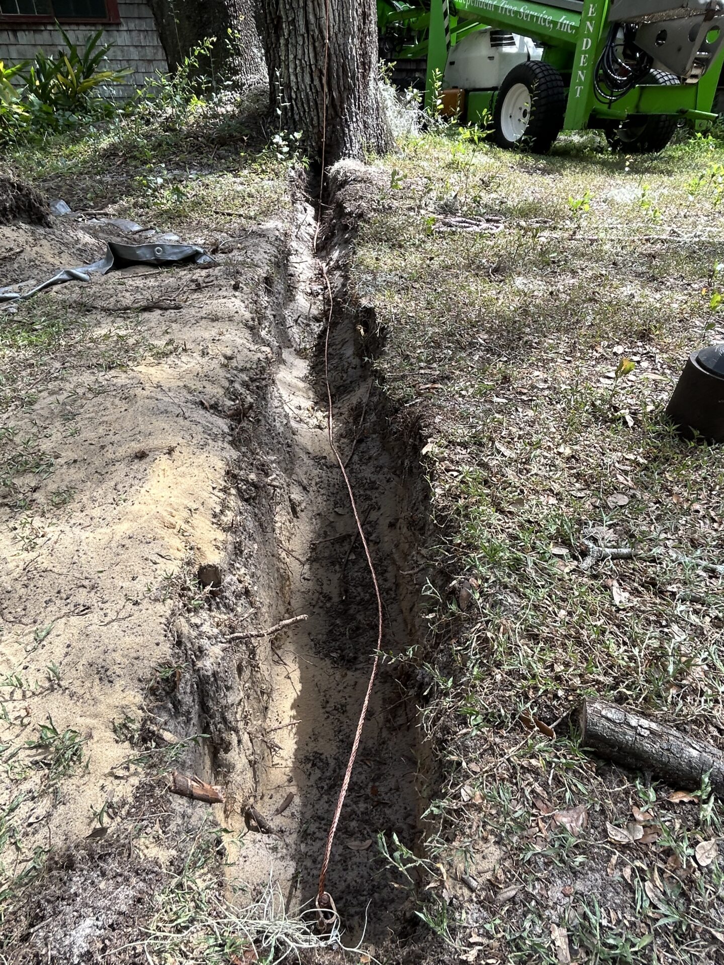 A trench in a wooded area with a cable inside; nearby is a green machine, possibly for construction or landscaping.