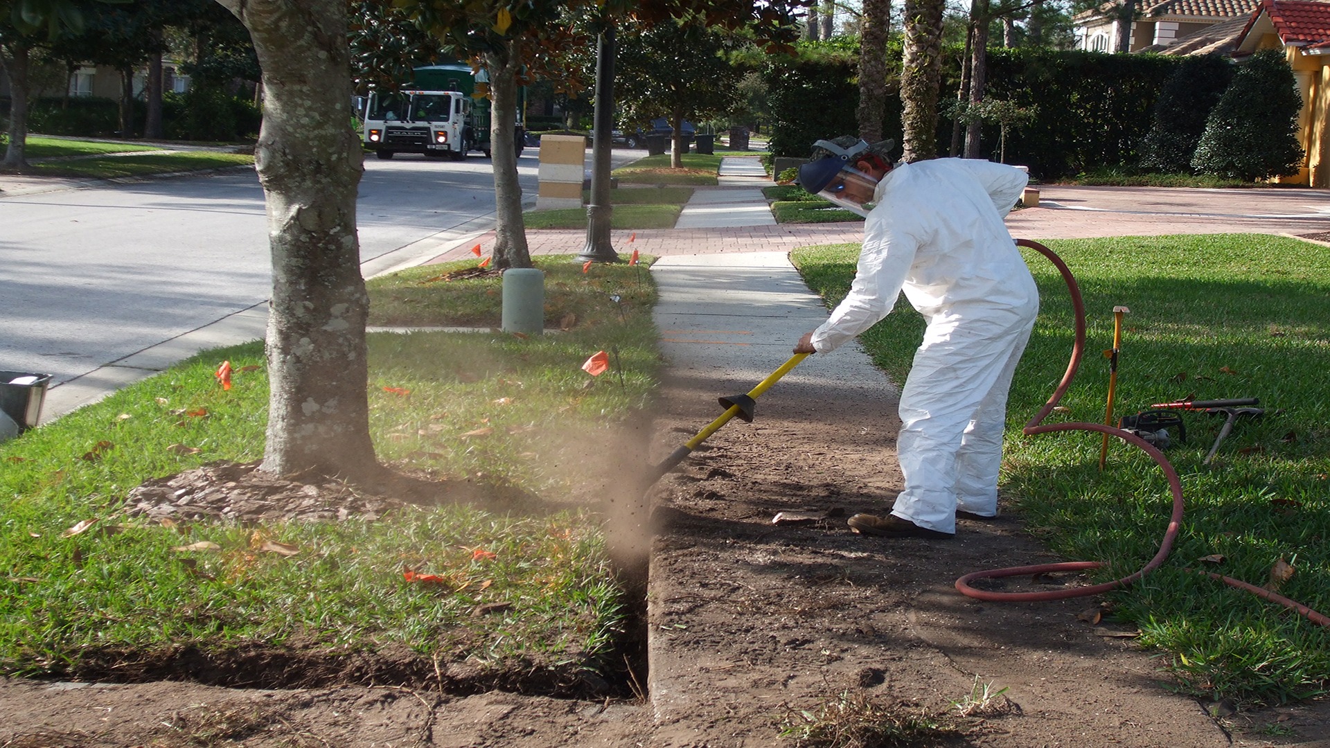 A person in protective gear uses equipment to edge a grassy sidewalk near trees on a residential street, with a vehicle in the background.