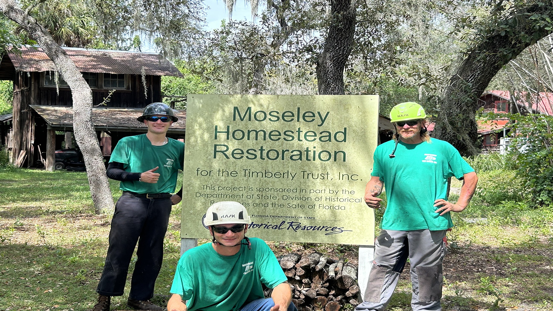 Three people in green shirts and helmets at Moseley Homestead Restoration sign, surrounded by trees and historic buildings in the background.