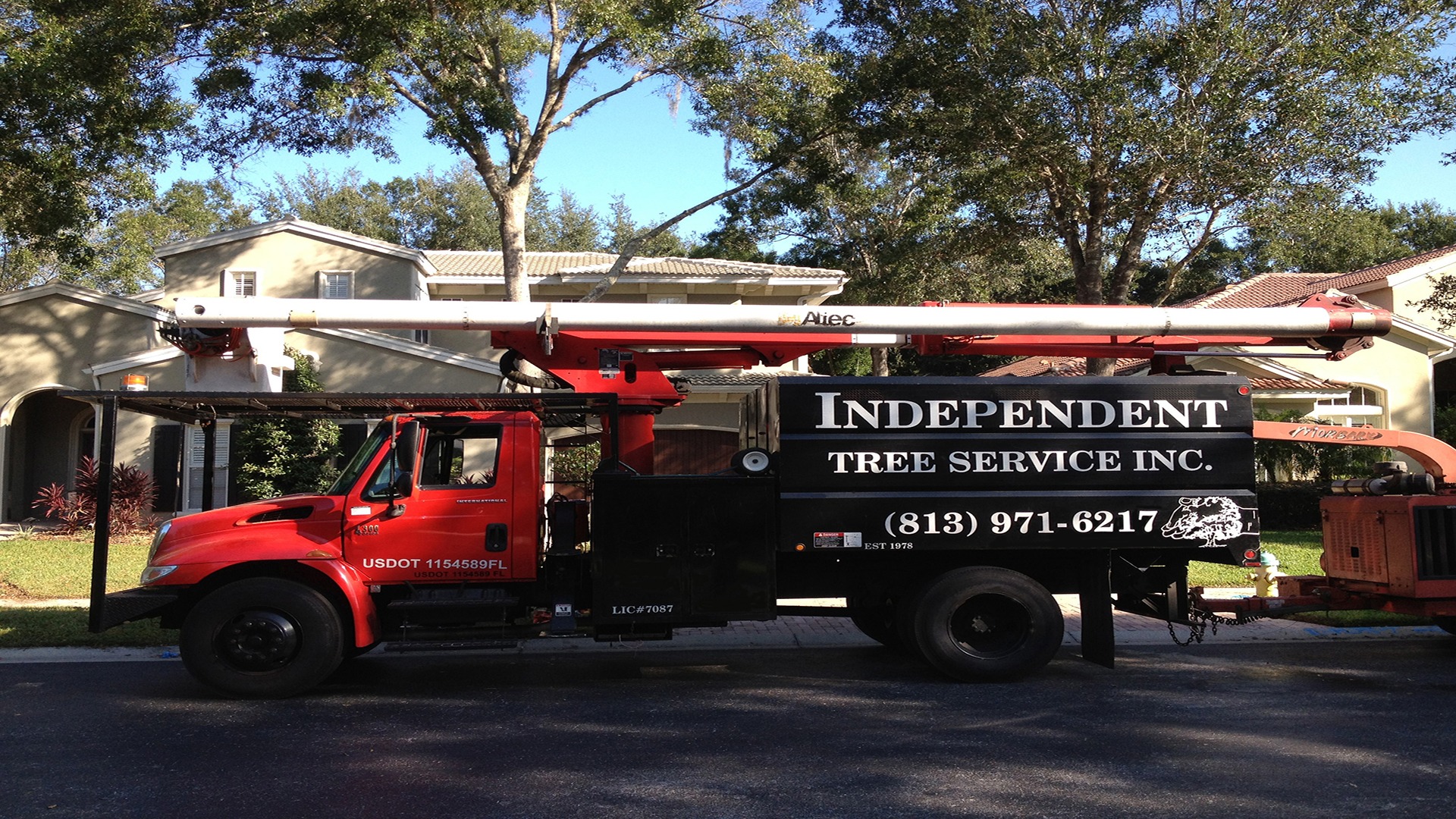 A red truck labeled "Independent Tree Service Inc." is parked on a residential street, surrounded by trees and houses in the background.